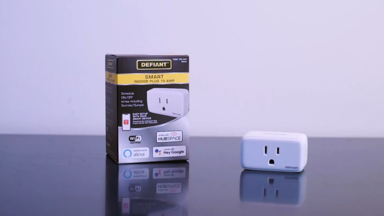 A white Defiant Indoor Smart Plug sits beside its product packaging on a glossy black surface.