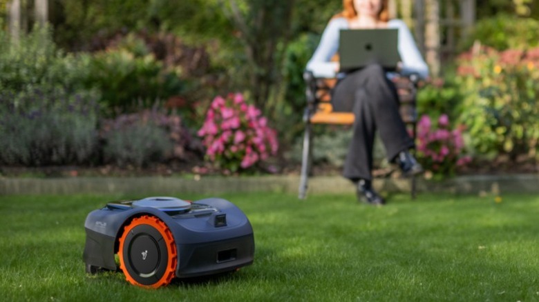 Robot lawn mower cutting grass while a woman works on laptop in the garden.
