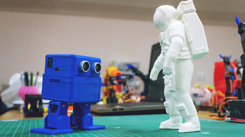 A 3D printed blue robot and white astronaut stand on a hobby mat facing each other.