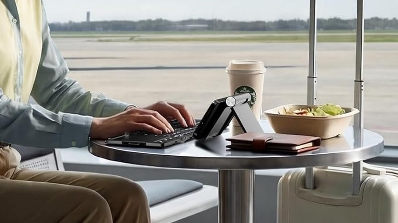 A pair of hands typing on the iClever BK08 Foldable Keyboard, placed on a circular table next to a coffee cup, a salad bowl, and a travel suitcase