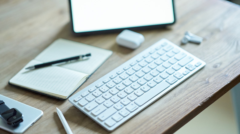 A Bluetooth keyboard placed on a wooden table with wireless earbuds outside their case, a stylus, a notebook, a smartwatch on top of a phone, and a tablet