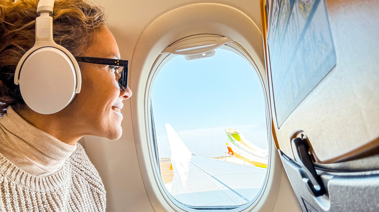A woman wearing noise-cancelling earphones and looking out a window on an airplane.