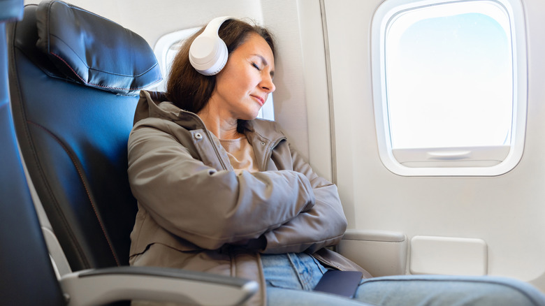 A woman sits in an airplane wearing headphones with her eyes closed.