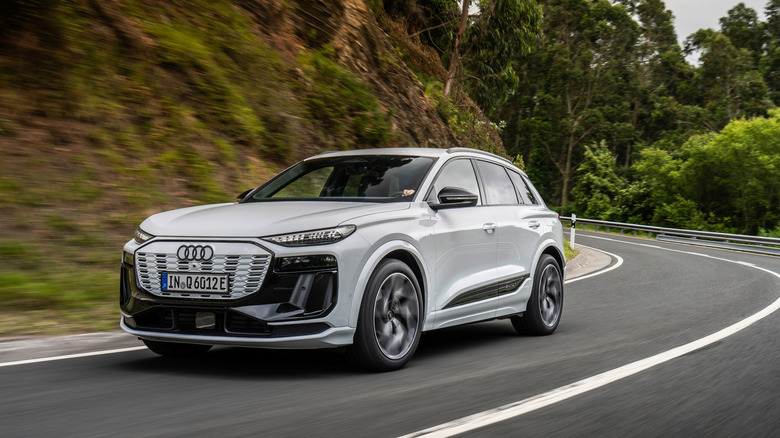 A light gray Audi Q6 e-tron driving along a downward road surrounded by green trees
