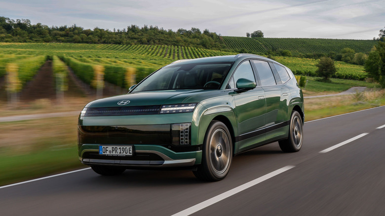 A green Hyundai Ioniq 9 driving on a paved road with a green field and trees in the background