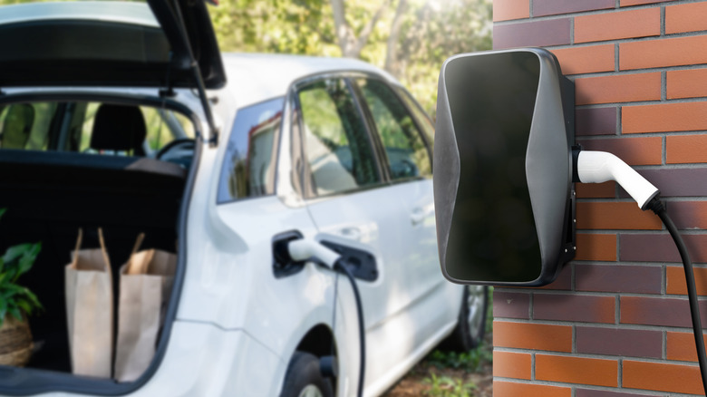A white hatchback SUV with paper bags in an open trunk charging beside a brick building