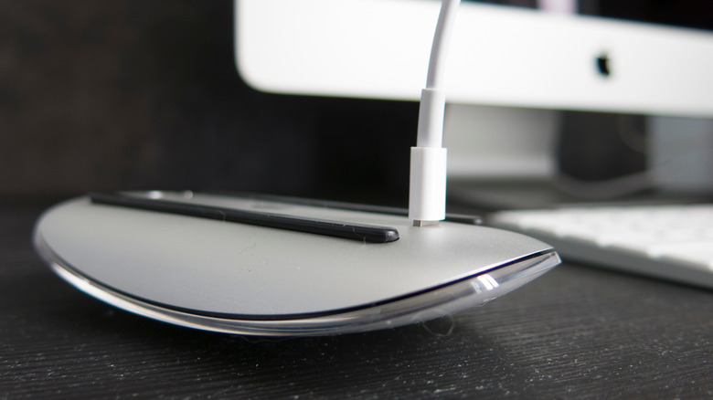 A Magic Mouse charging on a black desktop, the white cable sticking out of its underside.