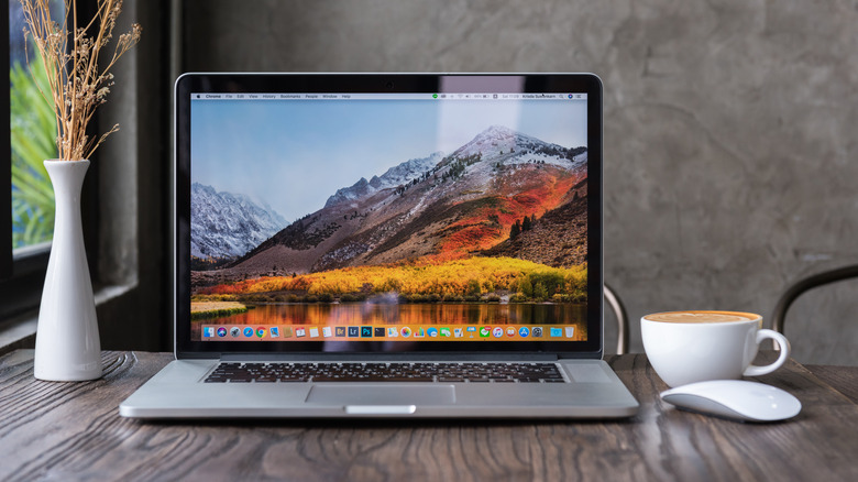A Macbook open, showing a desktop background of a mountain, on a gray wooden table next to a vase of dried flowers, a cup of coffee, and a white Magic Mouse.