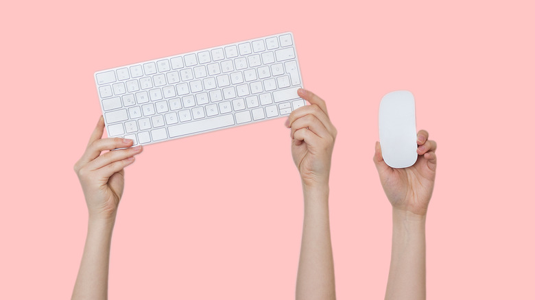 A set of three hands holding up a white Apple keyboard and magic mouse against a blush pink background.