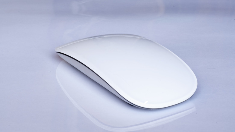 A white Apple Magic Mouse lying on a reflective white glass table.