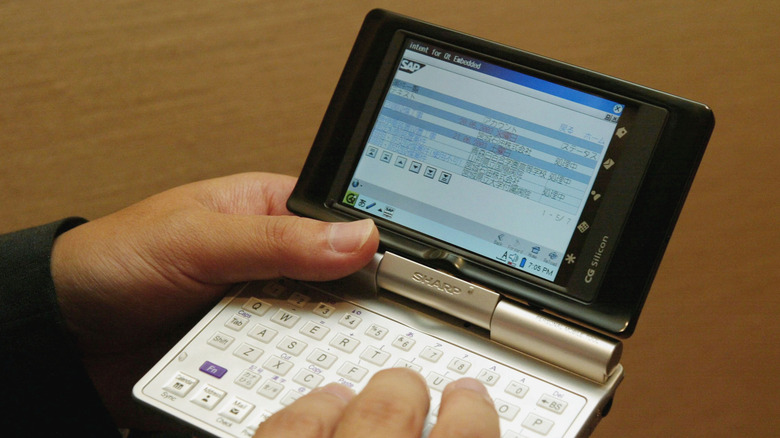 A person holds a Sharp clamshell PDA in both hands with the screen open.