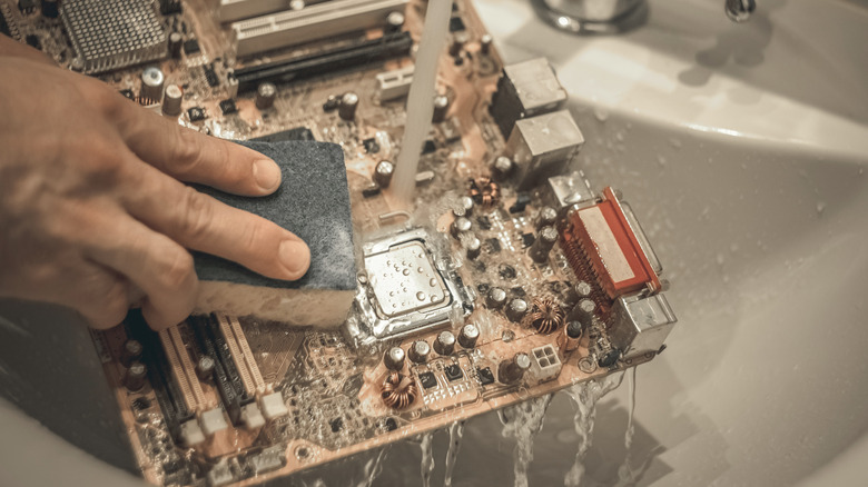 A close-up photo of someone washing a computer motherboard in a sink using running water and a sponge.