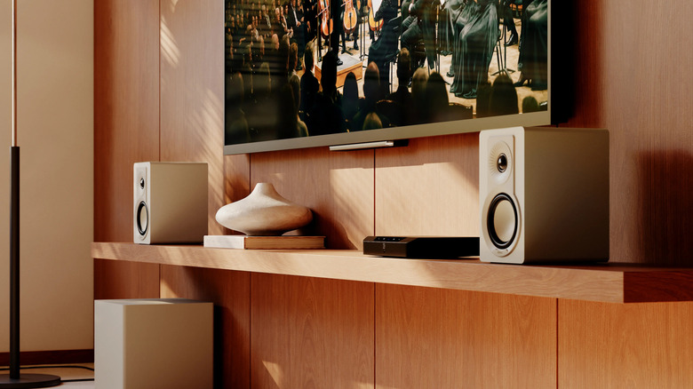 Dali Kupid speakers on a wooden shelf under a television mounted to a wall.