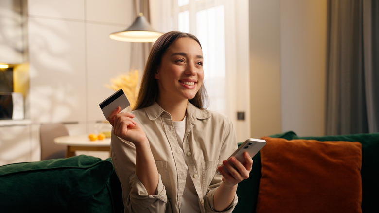 A woman sits on her couch holding her phone in one hand and her credit card in another.