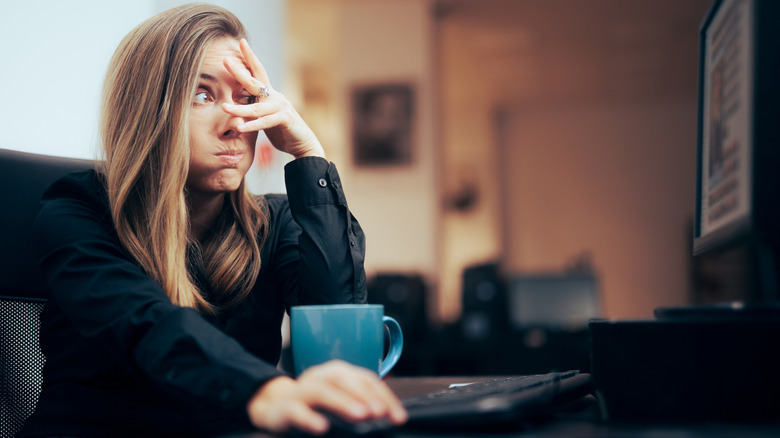 A woman sitting at her computer is partially covering her face as she has a strong reaction to the screen.