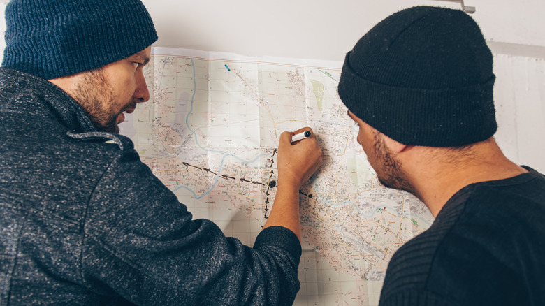 Two men in black clothes plot a course on a map against the wall.