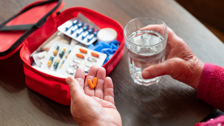 An old woman holds orange pills in one hand and a glass of water in the other. In front of her is a First Aid Kit.