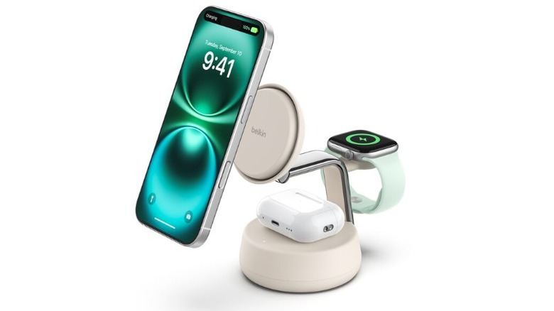 A product render of the Belkin UltraCharge Pro 3-in-1 Charging Dock in Sand color. An iPhone hovers over the charging pad, a pair of AirPods sit at the base, and an Apple Watch is charging at the back on a wireless puck.