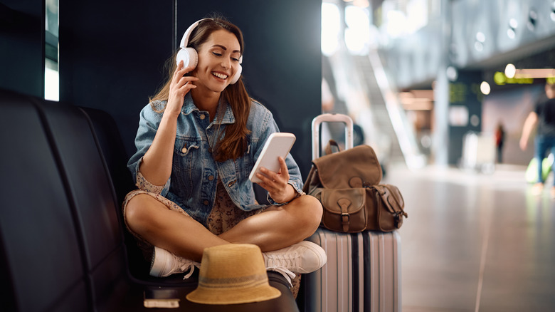 A woman sitting in and airport, wearing headphones and using her smartphone.