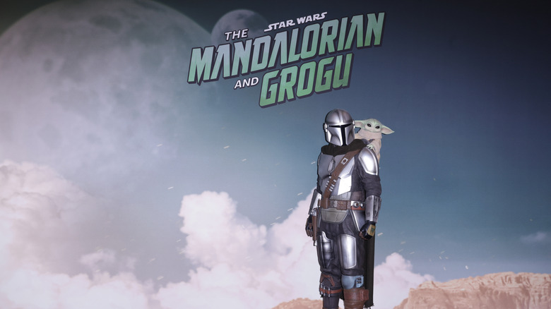 Promotional artwork for The Mandalorian and Grogu featuring the armored Mandalorian character with Grogu on his shoulder against a cloudy sci-fi backdrop.