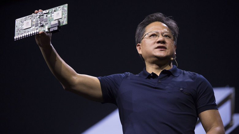 NVIDIA CEO Jensen Huang speaking on stage while holding up a GPU during a technology presentation.