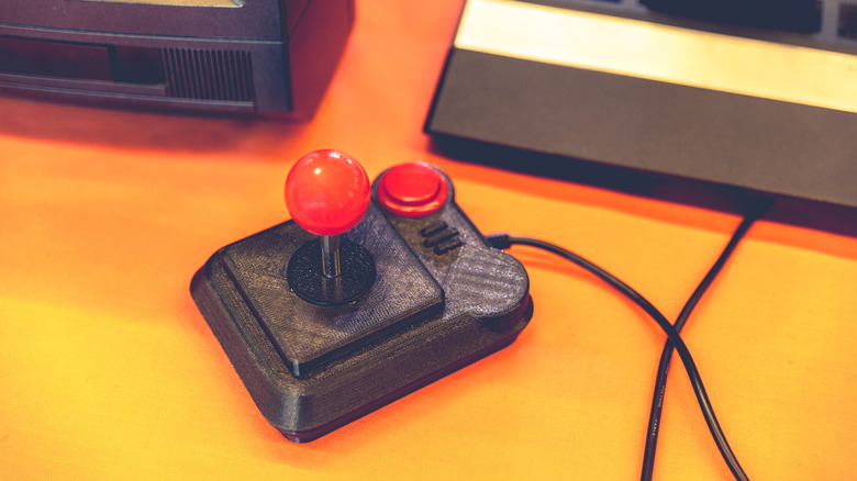 Close-up of a retro-style joystick controller with a red stick and button connected to a gaming system on an orange surface.