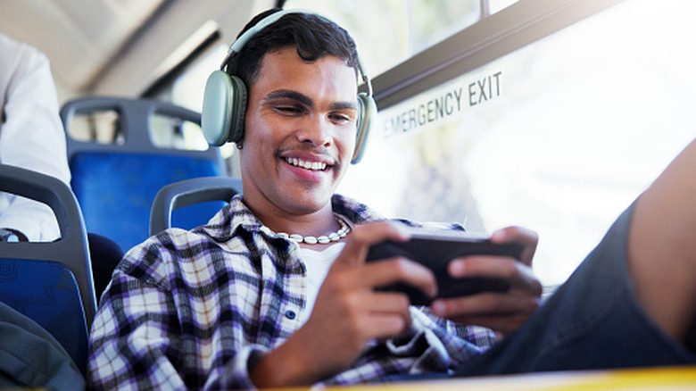 Young man wearing headphones and playing a game on a smartphone while riding on a bus.
