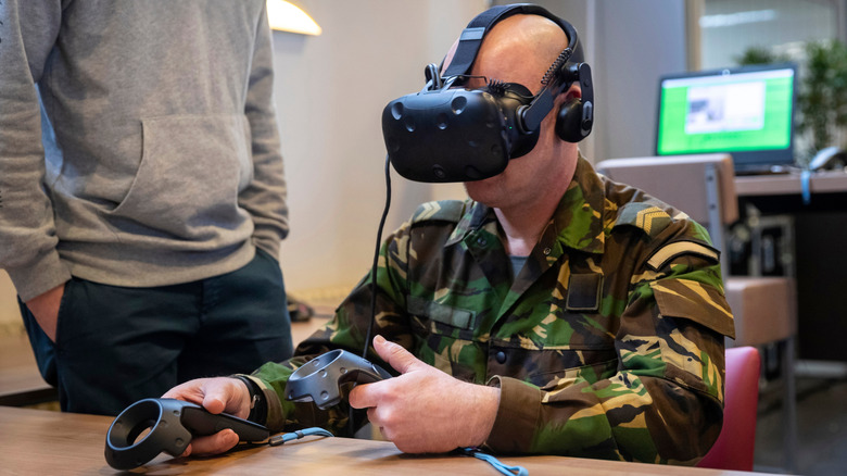 Person in military camouflage uniform using a virtual reality headset and handheld VR controllers while seated at a table indoors.
