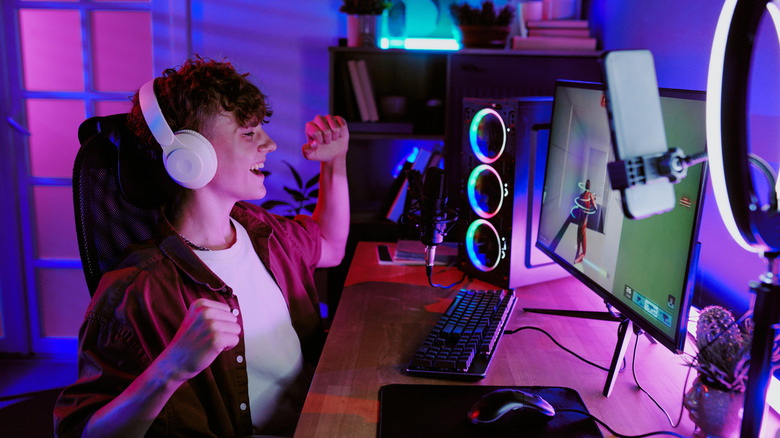 Young gamer or streamer celebrating at a desktop PC setup with colorful RGB lighting, headphones, microphone, and smartphone mounted in front of a ring light.