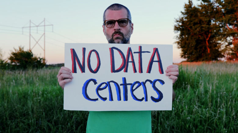 Man holding a large, hand-written "no data centers" sign in front of a field at dusk.