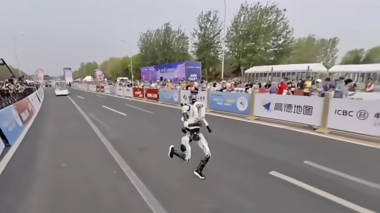 A white bipedal humanoid robot races down a track as spectators watch from the sidelines.