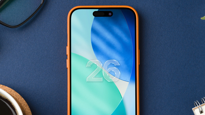 iOS 26 logo on iphone screen sitting on a dark blue table with other objects in background