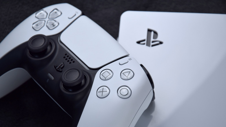 A white Sony Dual Shock Sense controller leans against a white PS5, both objects are against a black backdrop