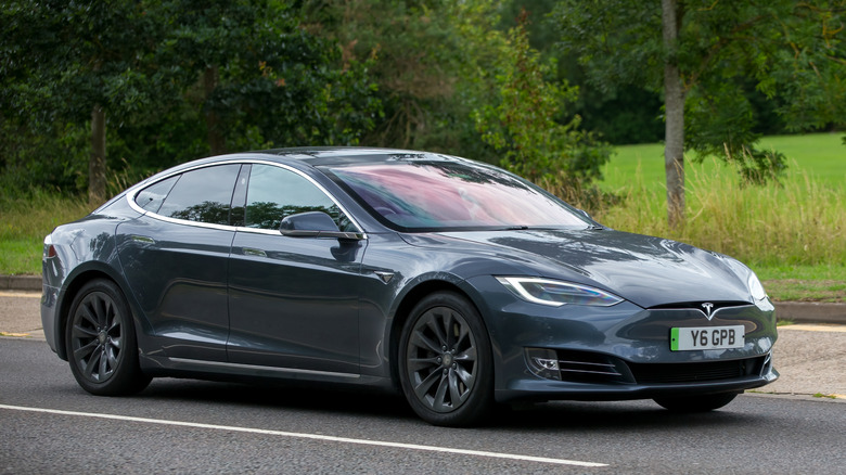 A black Tesla Model S as it sits on the road, grass and trees are behind the vehicle