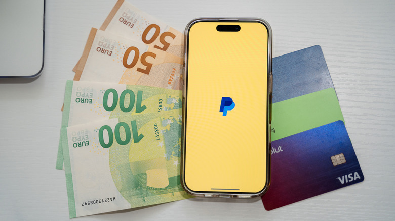 Aplicación PayPal en un iPhone junto a tarjetas de crédito y efectivo.