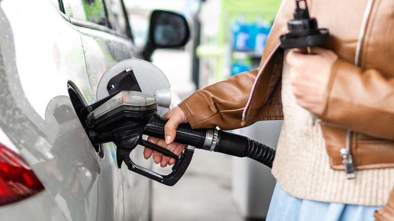 A individual in a brown leather jacket is using a fuel pump at a gas station to pump gas into a silver vehicle