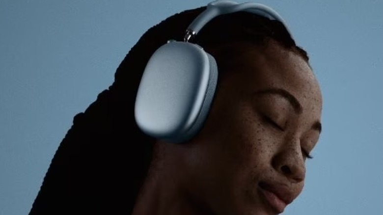 An individual wearing a pair of silver AirPods Max 2 on their head, the person is against a blue backdrop