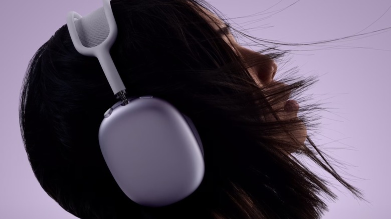 A pair of silver AirPods Max 2 adorned on an individual with black hair's head, the person is against a light purple backdrop