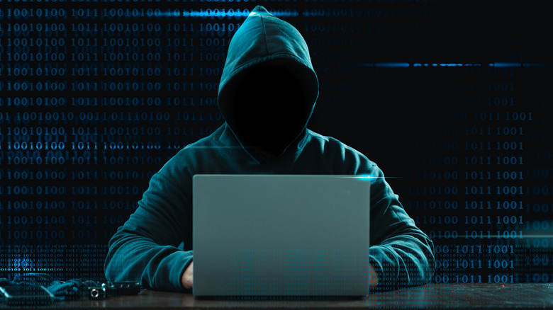 A hooded figure with their face shadowed using a laptop with lines of binary in the background