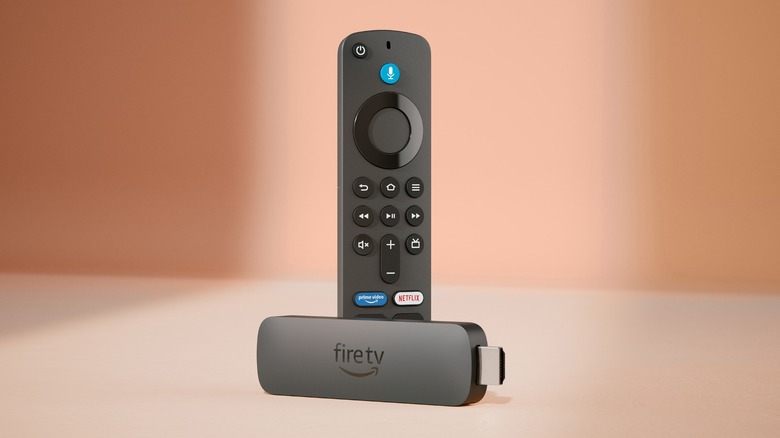 The Amazon Fire TV Stick 4K Plus device sitting in front of a vertical Fire TV remote, both objects are against a background of two different brownish hues