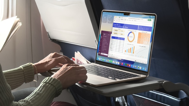 An individual seated on an AirPlane has a MacBook Air on a tray table, both of their hands are on the laptop as they work on documents