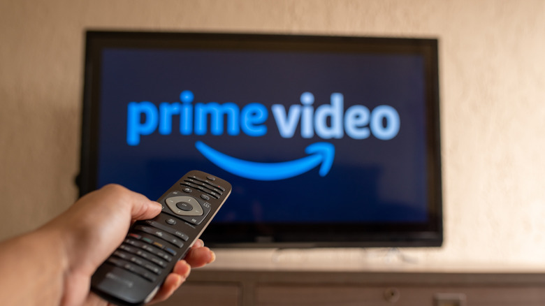 Close-up of a person holding a TV remote, with a blurred TV in the background feature Prime Video logo