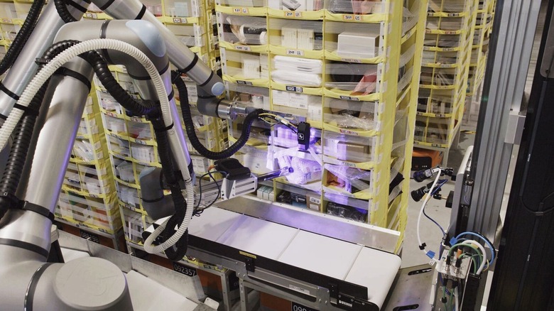 A silver Vulcan robot shines a purple light on a package it's pulled from a sorting cubby in order to scan it