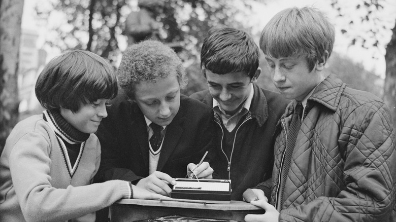 Four individuals standing outdoors next to each other, with one in the middle of the group having their hands on a Stylophone pocket synthesizer