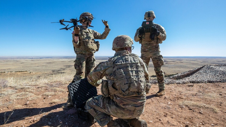 Three soldiers, one crouching in the foreground with an open case, another holding the drone up for launch, and a third staring at the control monitor, prepare to operate a drone in the desert landscapes around Fort Carson, Colorado