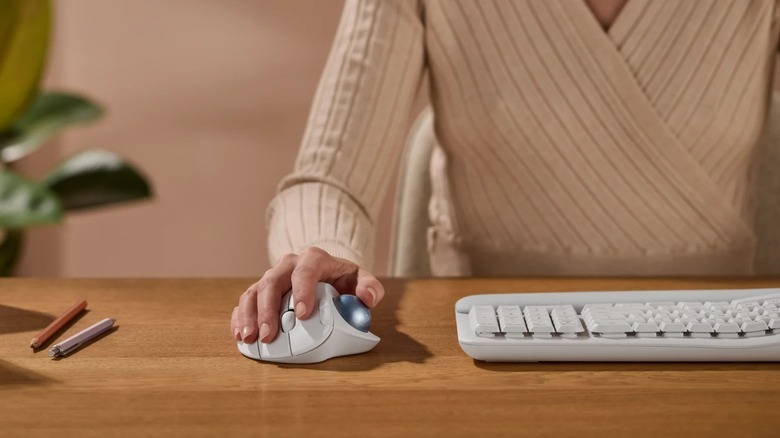 Hand holding Logitech Ergo M575S Wireless Trackball Mouse on desk with keyboard