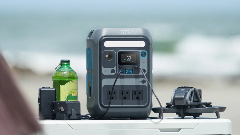 The Anker Solix C300 Portable Power Station sits between a drone and its accessories, there is also a green glass with a bottle cap filled with fluid sitting next to the charger
