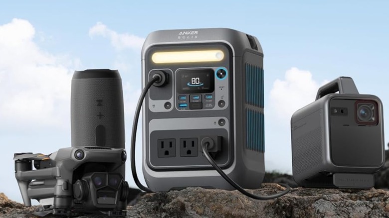 The Anker Solix C300 Portable Power Station sitting next to a projector, a sound bar, and what appears to be a drone, all of the items are sitting on a rock against a cloudy background