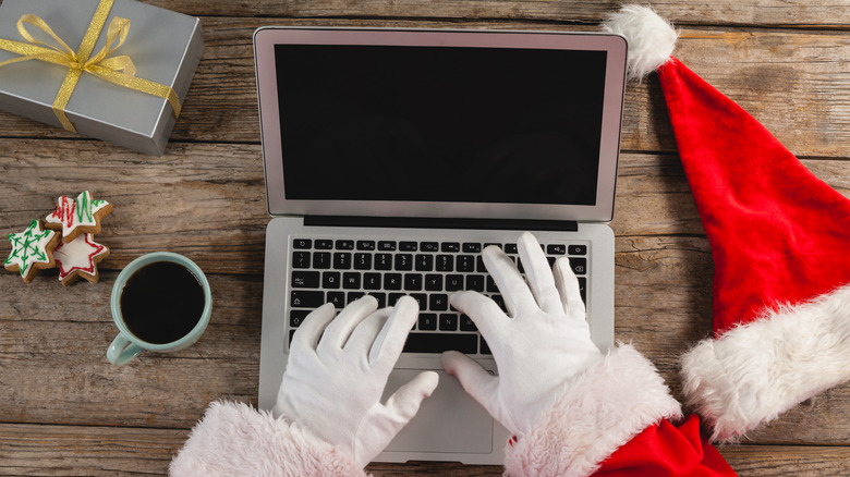 A photo of a person's hands in white gloves while they were a red suit with fluffy white trim about the sleeves, their hands are on a laptop keyboard and there is a red hat and a cup of coffee next to them