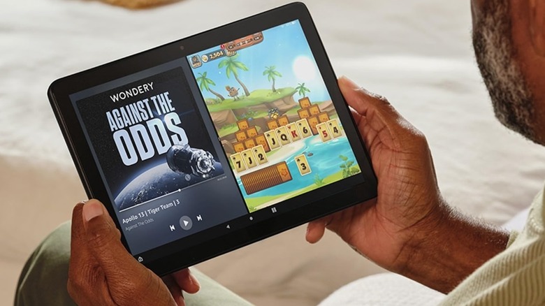 A behind-the-shoulder shot of an individual holding an Amazon Fire HD 10 horizontally in both their hands, the device is displaying a media player and some sort of card game